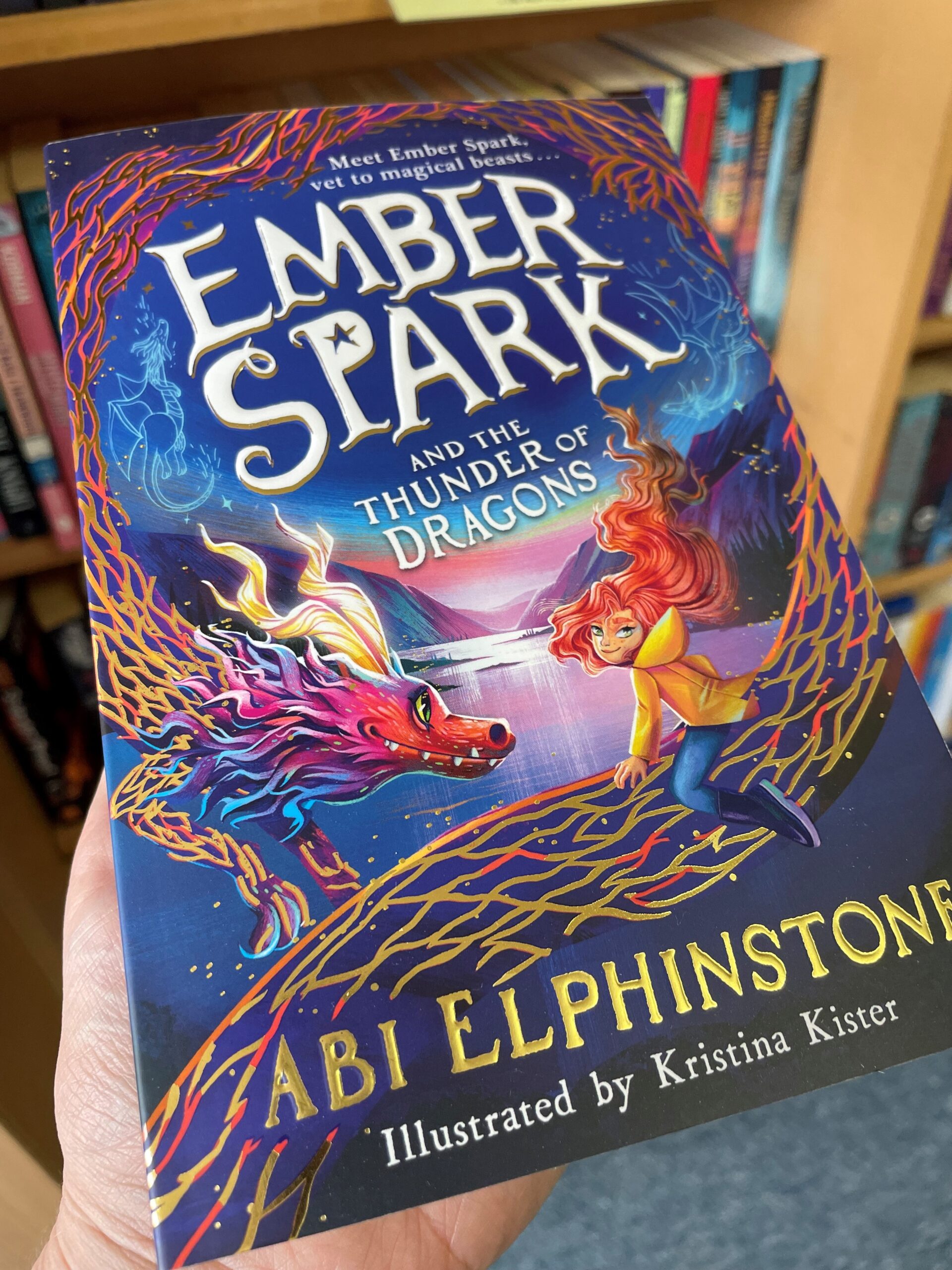 Book called Ember Spark and the Thunder of Dragons held by one hand in front of a bookcase.